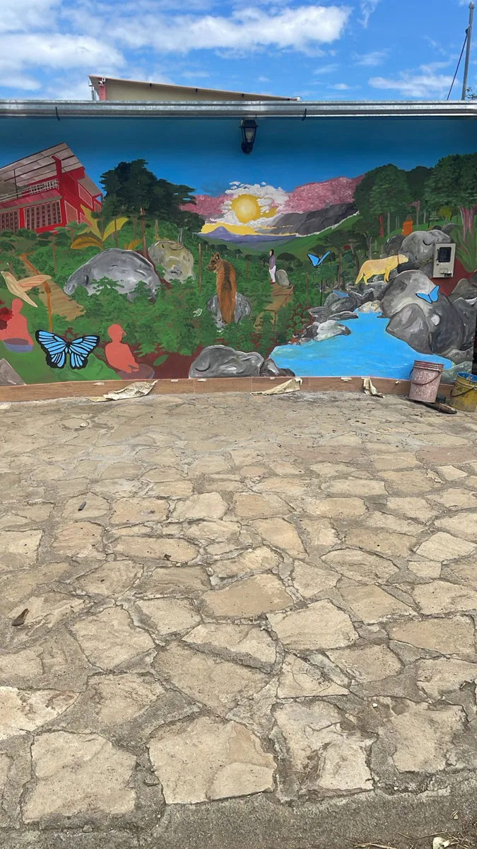 Mural en proceso con la casa de la finca, un río, árboles, animales y mariposas.